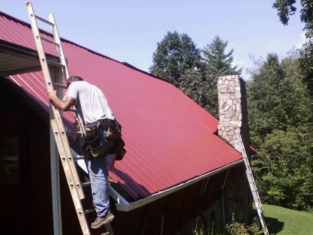 Roofer on a ladder beside a red metal roof and stone chimney — Soffit & Fascia Repair by Roofing Brothers in Leadore